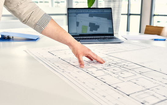 Auf einem Tisch sind Laptop und Bauplan; eine Hand zeigt auf den Bauplan