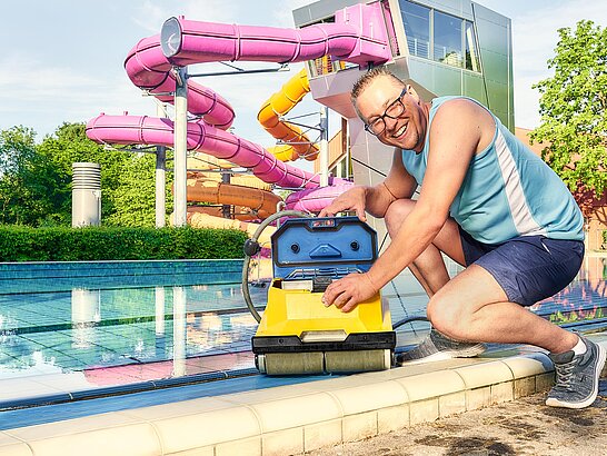Mann hockt an Beckenrand von Freibad und hält ein Reinigungsgerät in der Hand