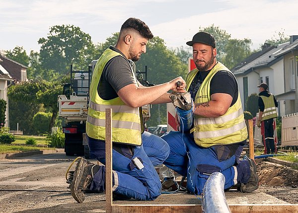 Zwei Männer im Blaumann und mit Warnwesten hocken an einer Baustelle; der eine Mann hält das Rohr der andere Schabt mit einer Kurbel etwas von dem Rohr ab