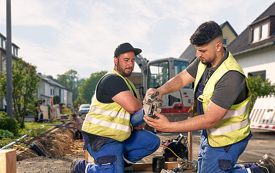 Zwei Männer im Blaumann hocken in einer Baustelle mit einem Bagger im Hintergrund; der ein Monteur hält ein Rohr der anderer schabt mit einem Schaber etwas von dem Anfang des Rohrs ab