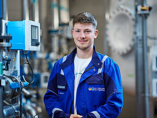 Mann mit blauer Jacke steht vor einer technischen Anlage und lächelt in die Kamera