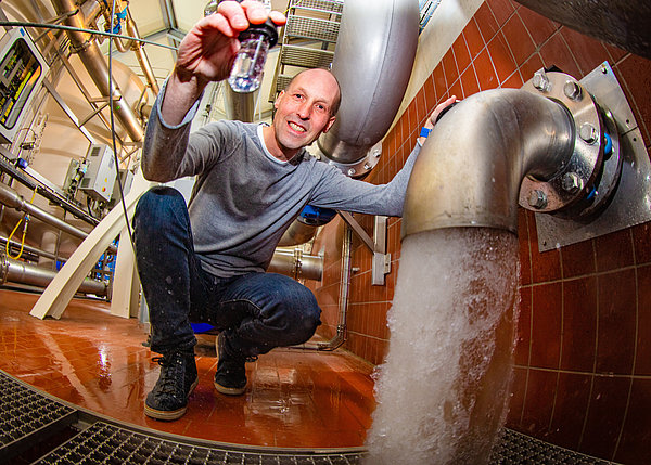 Ein Mann hockt in einem Wasserwerk vor einem laufenden Wasserstrahl und hält ein Glas mit Wasser hoch